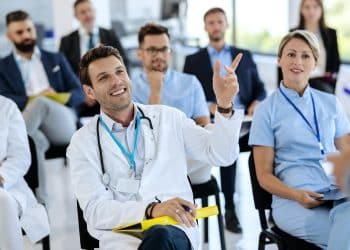 Profissionais de saúde de jaleco assistem a uma palestra, um deles com o braço levantado para participar