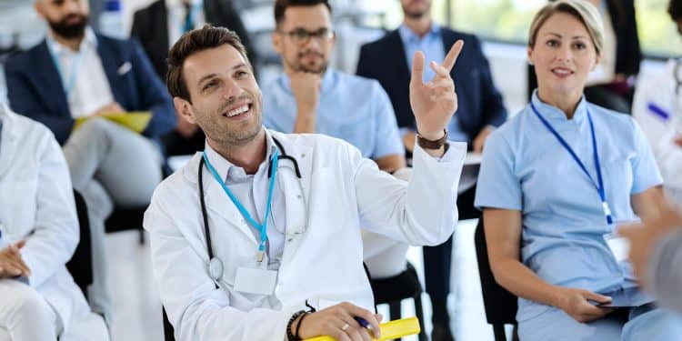 Profissionais de saúde de jaleco assistem a uma palestra, um deles com o braço levantado para participar