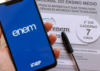 Mão segura celular com tela do ENEM ao lado de prova impressa e caneta preta.