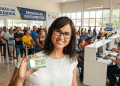 Mulher sorridente segurando a nova Carteira de Identidade Nacional durante mutirão de emissão de documentos, com fila de atendimento ao fundo