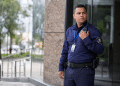 Segurança masculino com uniforme azul, crachá e rádio comunica-se de pé na entrada de prédio.