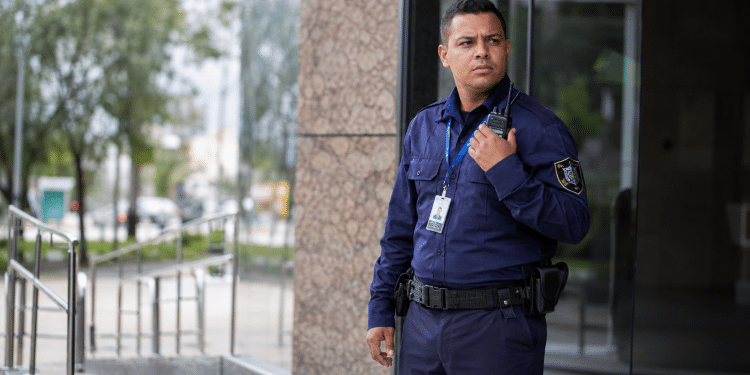 Segurança masculino com uniforme azul, crachá e rádio comunica-se de pé na entrada de prédio.