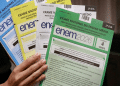 Mão segura quatro cadernos do ENEM 2026 em verde, branco, amarelo e azul com instruções visíveis, simbolizando os motivos que podem levar à eliminação no Enem.