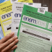 Mão segura quatro cadernos do ENEM 2026 em verde, branco, amarelo e azul com instruções visíveis, simbolizando os motivos que podem levar à eliminação no Enem.