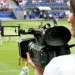 Cinegrafista em estádio de futebol registrando partida ao vivo, destacando atuação profissional no setor esportivo.