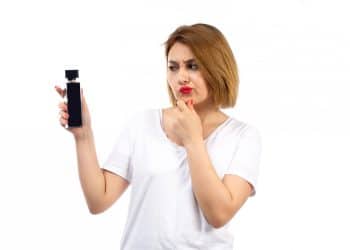 Mulher de camiseta branca observa frasco de perfume preto com expressão de dúvida