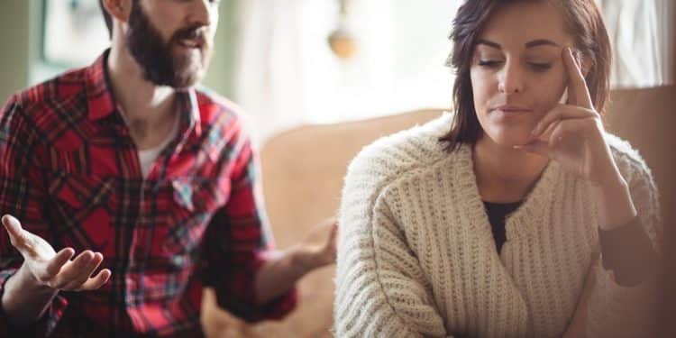 Mulher de suéter bege ignora homem de camisa xadrez vermelha que gesticula durante discussão no sofá, simbolizando frases que pessoas arrogantes costumam dizer com frequência.
