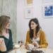 Duas mulheres sentadas à mesa de café, uma gesticulando enquanto a outra olha séria, parede decorada ao fundo.