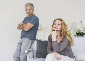 Homem de braços cruzados observa mulher sentada, ambos sérios em sala clara, simbolizando as frases que revelam que o respeito terminou.