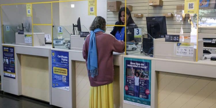Mulher idosa de cabelo grisalho e cacheado, casaco roxo e cachecol azul, sendo atendida por funcionária em agência dos Correios, simbolizando o funcionamento dos bancos nos jogos do Brasil.