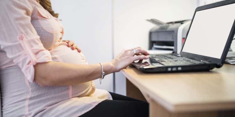 Mulher grávida sentada ao computador com uma mão na barriga e outra digitando, simbolizando o Projeto de Lei que propõe participação remota e segunda chamada para gestantes em concursos públicos.