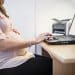 Mulher grávida sentada ao computador com uma mão na barriga e outra digitando, simbolizando o Projeto de Lei que propõe participação remota e segunda chamada para gestantes em concursos públicos.