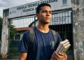 Jovem estudante com caderno e livros na mão e mochila nas costas em frente à escola, representando as vagas em cursos técnicos do programa Jovens do Amanhã