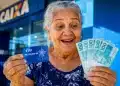 Idosa brasileira sorrindo segura cartão de CPF e notas de real em frente a uma agência bancária, representando os pagamentos de junho.