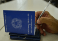 Mão masculina assinando Carteira de Trabalho azul com caneta em superfície branca, simbolizando o pagamento do lote extra do PIS/Pasep.