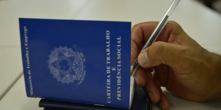 Mão masculina assinando Carteira de Trabalho azul com caneta em superfície branca, simbolizando o pagamento do lote extra do PIS/Pasep.