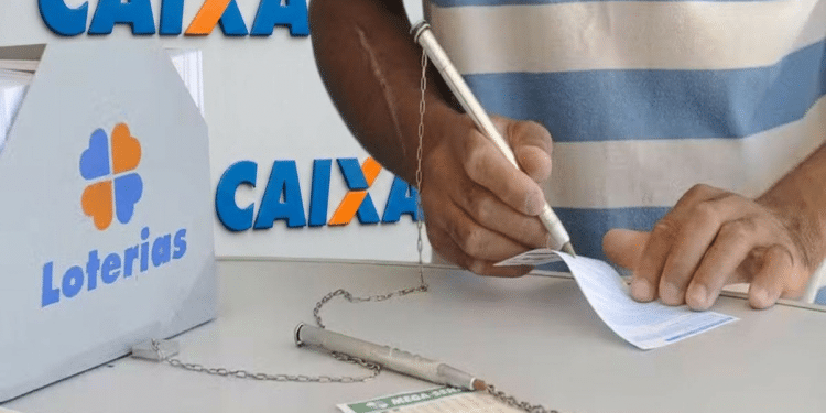 Pessoa de pele parda preenche bilhete da Mega-Sena ao lado de urna da Loterias Caixa, usando caneta presa por corrente, simbolizando a mudança de horário das loterias CAIXA.