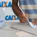 Pessoa de pele parda preenche bilhete da Mega-Sena ao lado de urna da Loterias Caixa, usando caneta presa por corrente, simbolizando a mudança de horário das loterias CAIXA.