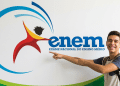 Jovem sorridente aponta para o logotipo do Enem fixado em parede branca
