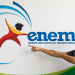 Jovem sorridente aponta para o logotipo do Enem fixado em parede branca