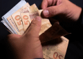 Pessoa conta notas de cinquenta reais segurando cédulas brasileiras em fundo escuro, simbolizando o pagamento de R$ 2.821 nos dias 1, 2 e 3 de julho.