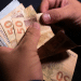 Pessoa conta notas de cinquenta reais segurando cédulas brasileiras em fundo escuro, simbolizando o pagamento de R$ 2.821 nos dias 1, 2 e 3 de julho.