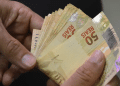 Mãos contando várias notas de cinquenta reais em fundo escuro, simbolizando o pagamento de R$ 1.621 do INSS no dia 29 de junho.