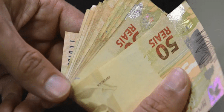 Mãos contando várias notas de cinquenta reais em fundo escuro, simbolizando o pagamento de R$ 1.621 do INSS no dia 29 de junho.