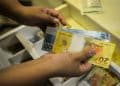 Mãos seguram notas de 20 e 100 reais sobre gaveta de caixa registradora aberta com divisórias, simbolizando o pagamento do teto do INSS em 3 e 5 de junho.