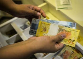 Mãos seguram notas de 20 e 100 reais sobre gaveta de caixa registradora aberta com divisórias, simbolizando o pagamento do teto do INSS em 3 e 5 de junho.