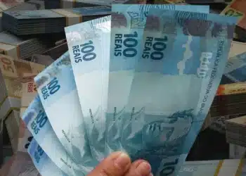 Mão segurando cédulas de R$ 100 em destaque, simbolizando a liberação de pagamento de R$ 2.621 em 1º de julho.