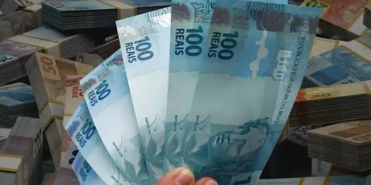 Mão segurando cédulas de R$ 100 em destaque, simbolizando a liberação de pagamento de R$ 2.621 em 1º de julho.