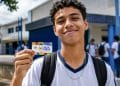 Jovem estudante uniformizado segura o cartão do Pé-de-Meia em frente à escola, representando os contemplados na nova rodada de pagamentos.