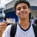 Jovem estudante uniformizado segura o cartão do Pé-de-Meia em frente à escola, representando os contemplados na nova rodada de pagamentos.