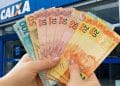 Mãos seguram cédulas diante de agência da Caixa, representando o pagamento do Pé-de-Meia de junho