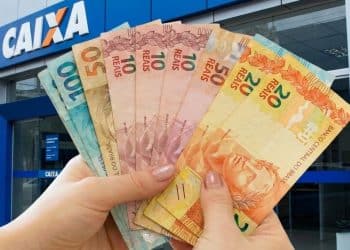 Mãos seguram cédulas diante de agência da Caixa, representando o pagamento do Pé-de-Meia de junho