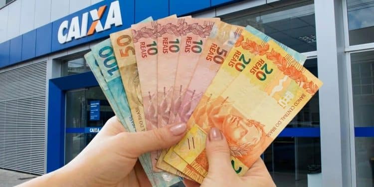 Mãos seguram cédulas diante de agência da Caixa, representando o pagamento do Pé-de-Meia de junho
