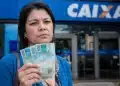 Mulher segura notas de 100 reais em frente a uma agência da Caixa, representando o pagamento tardio de R$ 1.621 para CPFs com finais de 0 a 9.