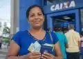 Mulher segura cédulas de reais e documentos em frente a uma agência da Caixa, representando o pagamento unificado do Bolsa Família em junho.
