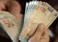 Mão segurando cédulas de real em destaque, representando pagamentos de R$ 946, R$ 1.216 e R$ 1.486 confirmados pela CAIXA.
