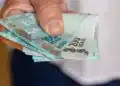 Mão segurando cédulas de real, representando pagamentos do INSS, abono PIS/Pasep e ressarcimentos liberados antes de julho.