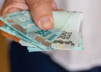 Mão segurando cédulas de real, representando pagamentos do INSS, abono PIS/Pasep e ressarcimentos liberados antes de julho.