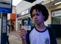 Estudante de uniforme observa o cartão do Pé-de-Meia em frente a uma agência da CAIXA, representando as dúvidas sobre o saque dos incentivos do programa.