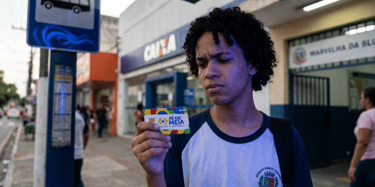 Estudante de uniforme observa o cartão do Pé-de-Meia em frente a uma agência da CAIXA, representando as dúvidas sobre o saque dos incentivos do programa.