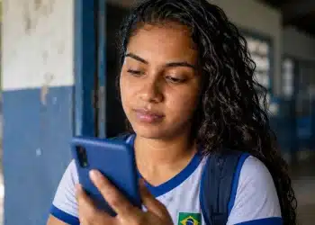 Estudante brasileira, com uniforme escolar, consultando a Conta CAIXA Tem no celular, com cédulas de dinheiro na mão, na entrada da escola.