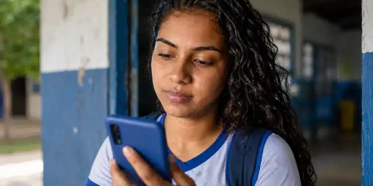Estudante brasileira, com uniforme escolar, consultando a Conta CAIXA Tem no celular, com cédulas de dinheiro na mão, na entrada da escola.