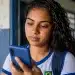 Estudante brasileira, com uniforme escolar, consultando a Conta CAIXA Tem no celular, com cédulas de dinheiro na mão, na entrada da escola.