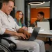 Profissional com deficiência em cadeira de rodas utilizando notebook em ambiente corporativo moderno, com colegas ao fundo em reunião colaborativa.