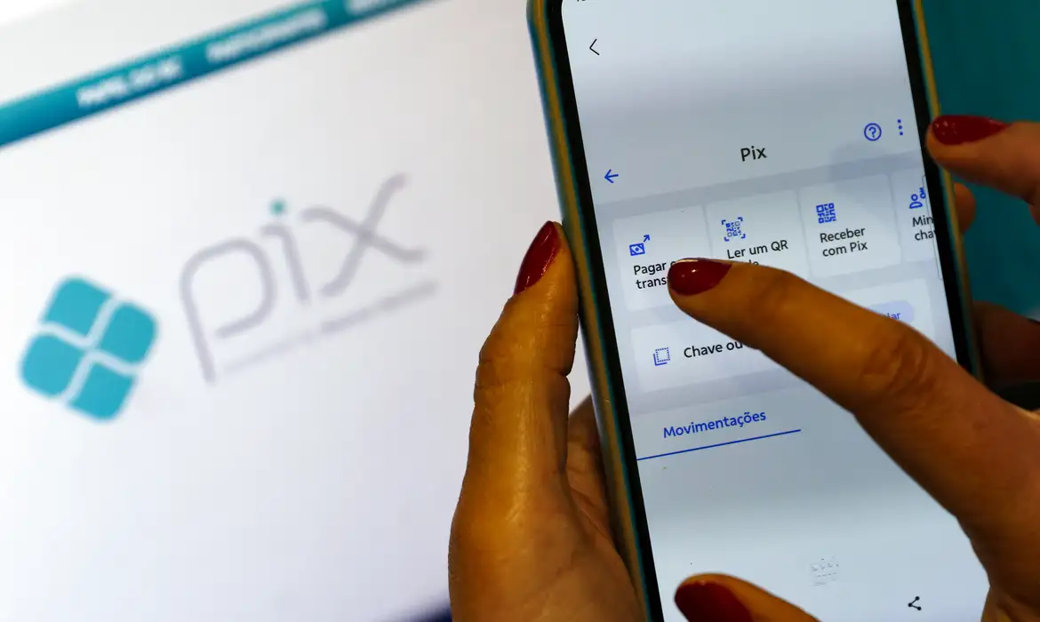Mão feminina com unhas vermelhas usando smartphone no app Pix com tela mostrando opções de pagamento, simbolizando o Pix de até R$ 1.000 referente à restituição automática do Imposto de Renda.
