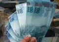 Mão segurando notas de 100 reais com pilhas de dinheiro ao fundo, representando pagamento do Pix automático para CPF aprovado.